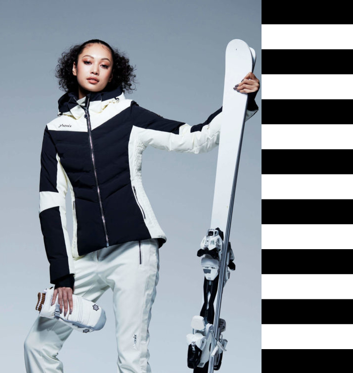 Model mit schwarz-weißer Skijacke, weißer Hose und Skiern in der Hand, Funktionsbekleidung für den Wintersport