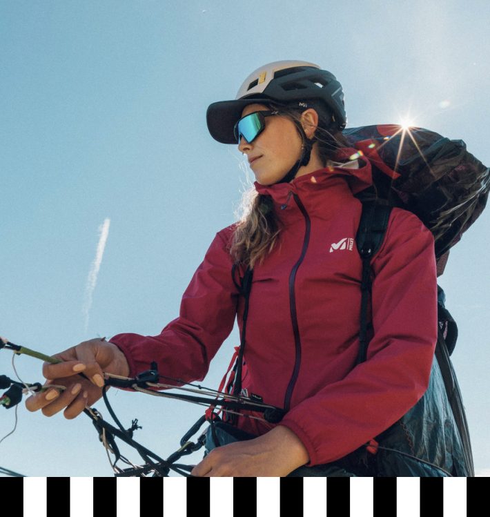 Frau mit einer rubinroten Hardshell-Jacke von Millet, Helm und Sonnenbrille, bereit für Outdoor-Aktivitäten in den Bergen, Blick auf …