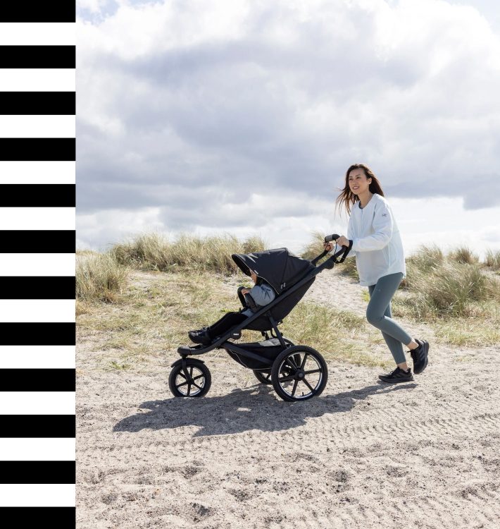 Frau, die einen Thule-Trekking-Kinderwagen auf einem sandigen Weg schiebt, ideal für jedes Gelände, im Freien, bewölkter Himmel