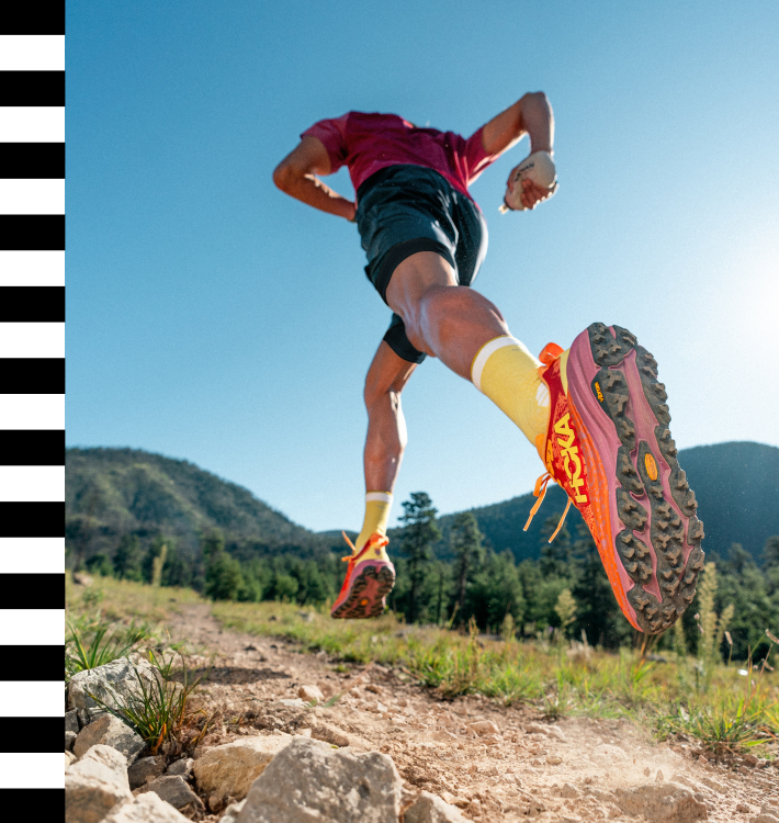 Läufer auf einem Feldweg mit orangefarbenen Trailrunning-Schuhen, Blick von unten, Berglandschaft und blauer Himmel
